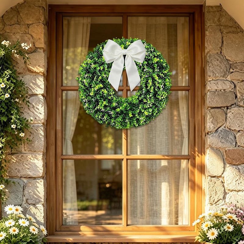 A green boxwood wreath with a white bow on a sunlit doorway